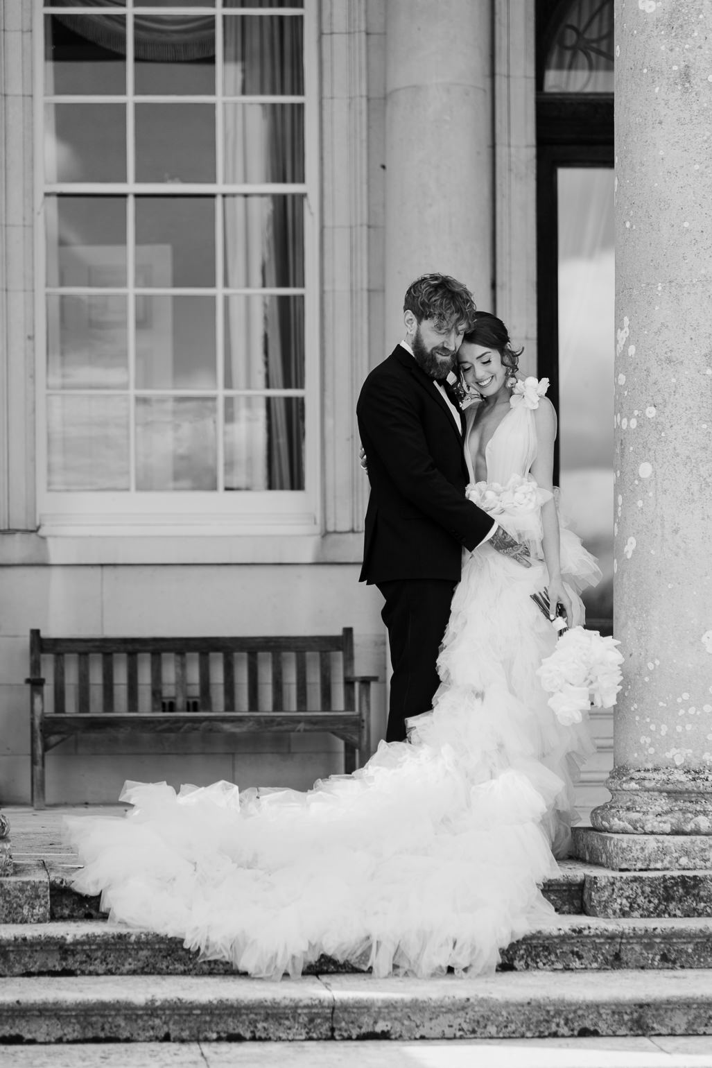 Bride in white ruffled Millia London wedding dress with groom in black tie standing on the steps of Stansted House, Stansted Park
