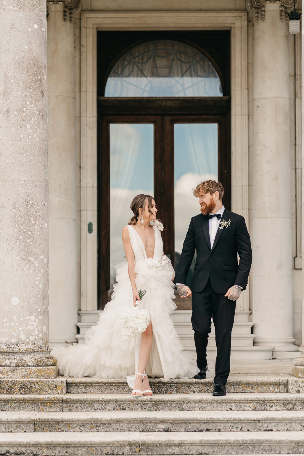 Bride in white ruffled Millia London wedding dress with groom in black tie standing on the steps of Stansted House