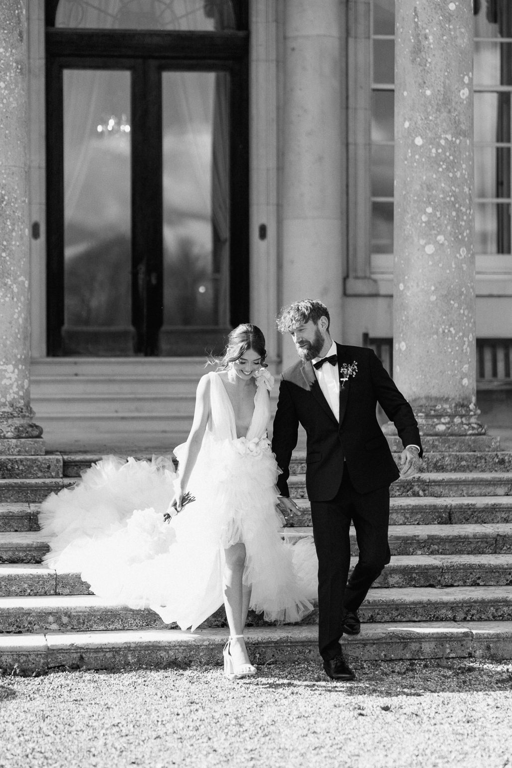 Bride in white ruffled Millia London wedding dress with groom in black tie standing on the steps of Stansted House